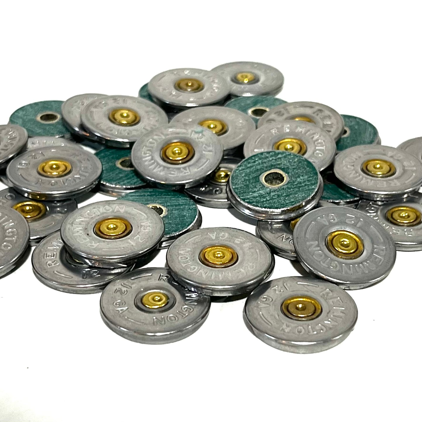 Large pile of Remington 12 gauge shotgun shell slices scattered on white background, showing a mix of silver headstamp faces and green hull-side faces with drilled center holes