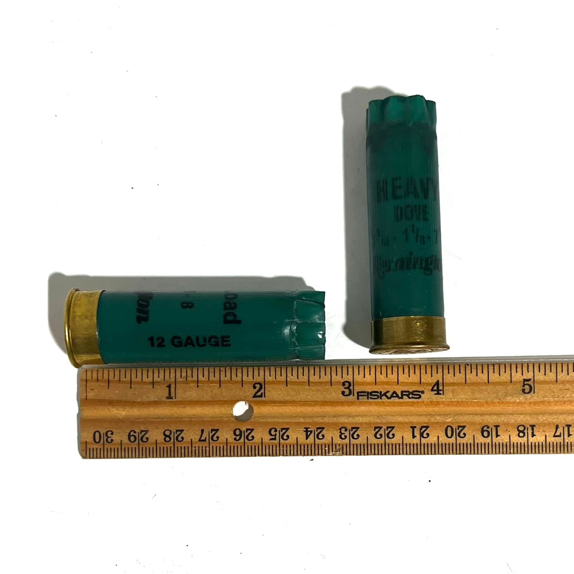 Two dark green Remington 12 gauge empty shotgun shells laid next to a wooden ruler for size comparison