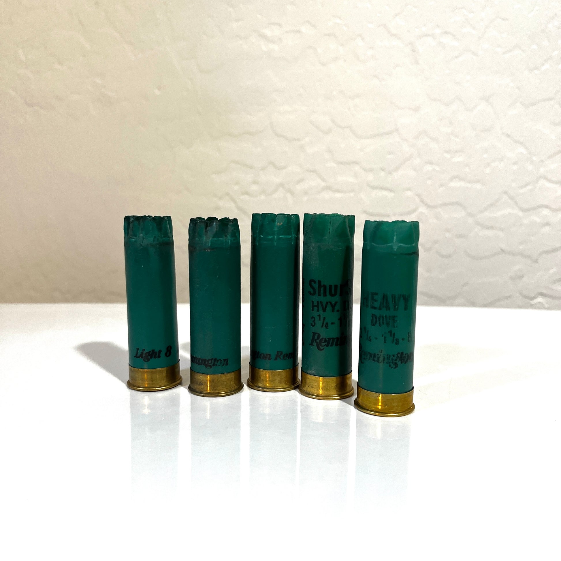Five dark green Remington 12 gauge empty shotgun shells with gold bases standing on a white surface