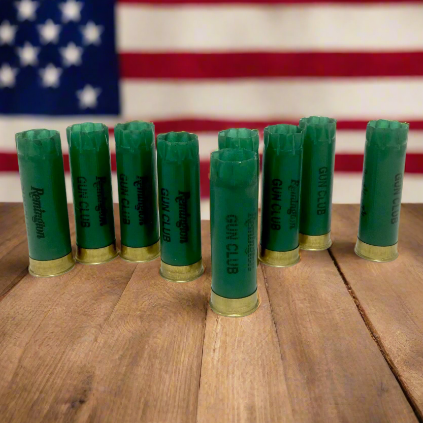 Nine green Remington Gun Club 12 gauge empty shotgun shells with gold bases standing on a wood surface with an American flag background