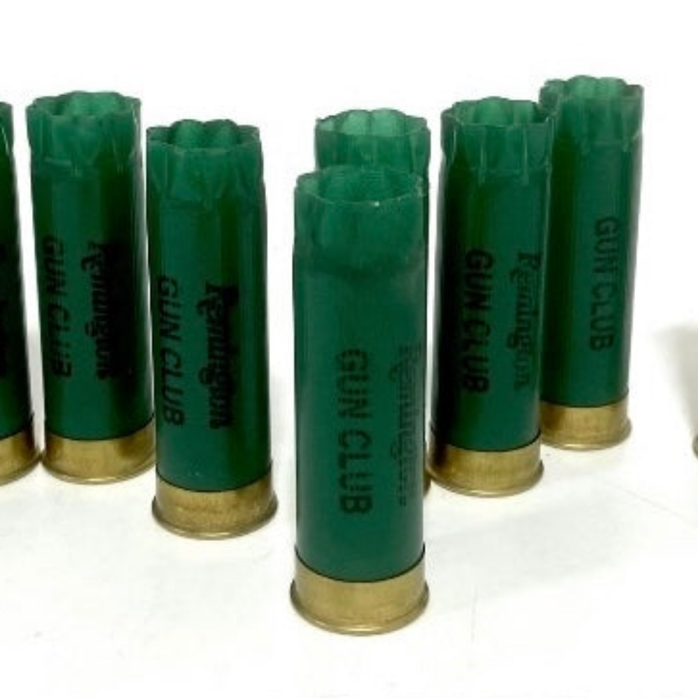Seven green Remington Gun Club 12 gauge empty shotgun shells with gold bases standing on a white background