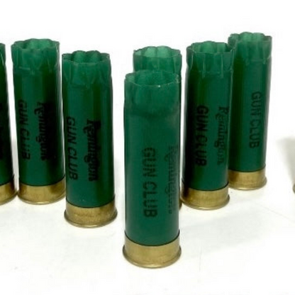 Seven green Remington Gun Club 12 gauge empty shotgun shells with gold bases standing on a white background