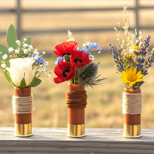 Three rose gold 12 gauge blank shotgun shells holding colorful flowers on wooden surface, rustic wedding boutonnieres.
