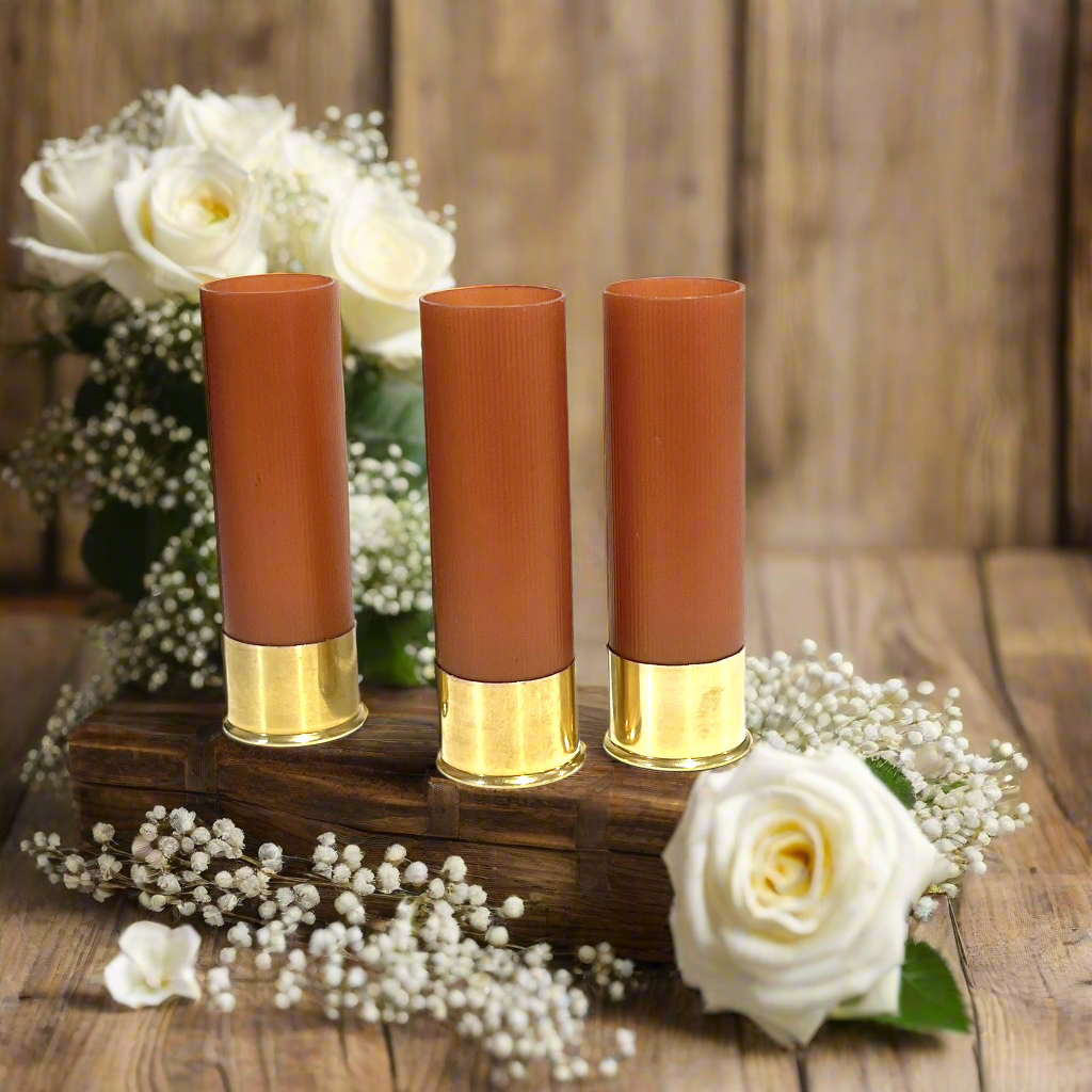 Three unmarked rose gold shotgun shells with high brass bottoms on wooden tray with white roses and baby's breath.
