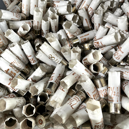 Overhead view of a dense pile of scattered white Gamebore 12 gauge fired shotgun shells with silver bases and various load markings.
