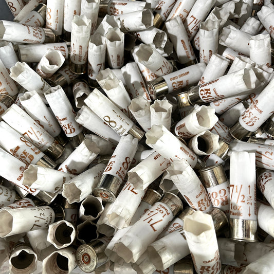 Overhead view of a dense pile of scattered white Gamebore 12 gauge fired shotgun shells with silver bases and various load markings.
