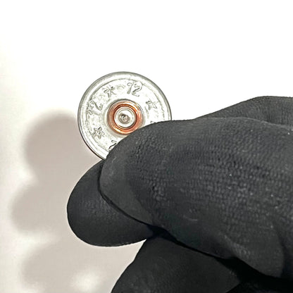 Close-up of a single silver shotgun shell slice with 12 and star stamped design and copper primer, held between black-gloved fingers