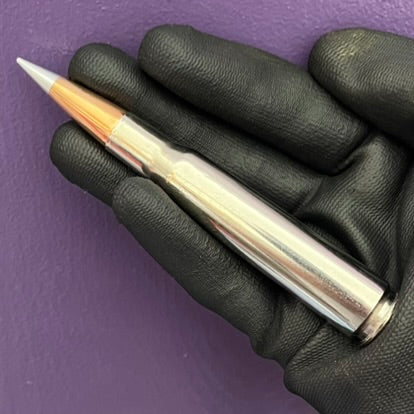Black gloved hand holding silver nickel 50 cal BMG dummy round with copper Hornady A-Max bullet tip on purple background.
