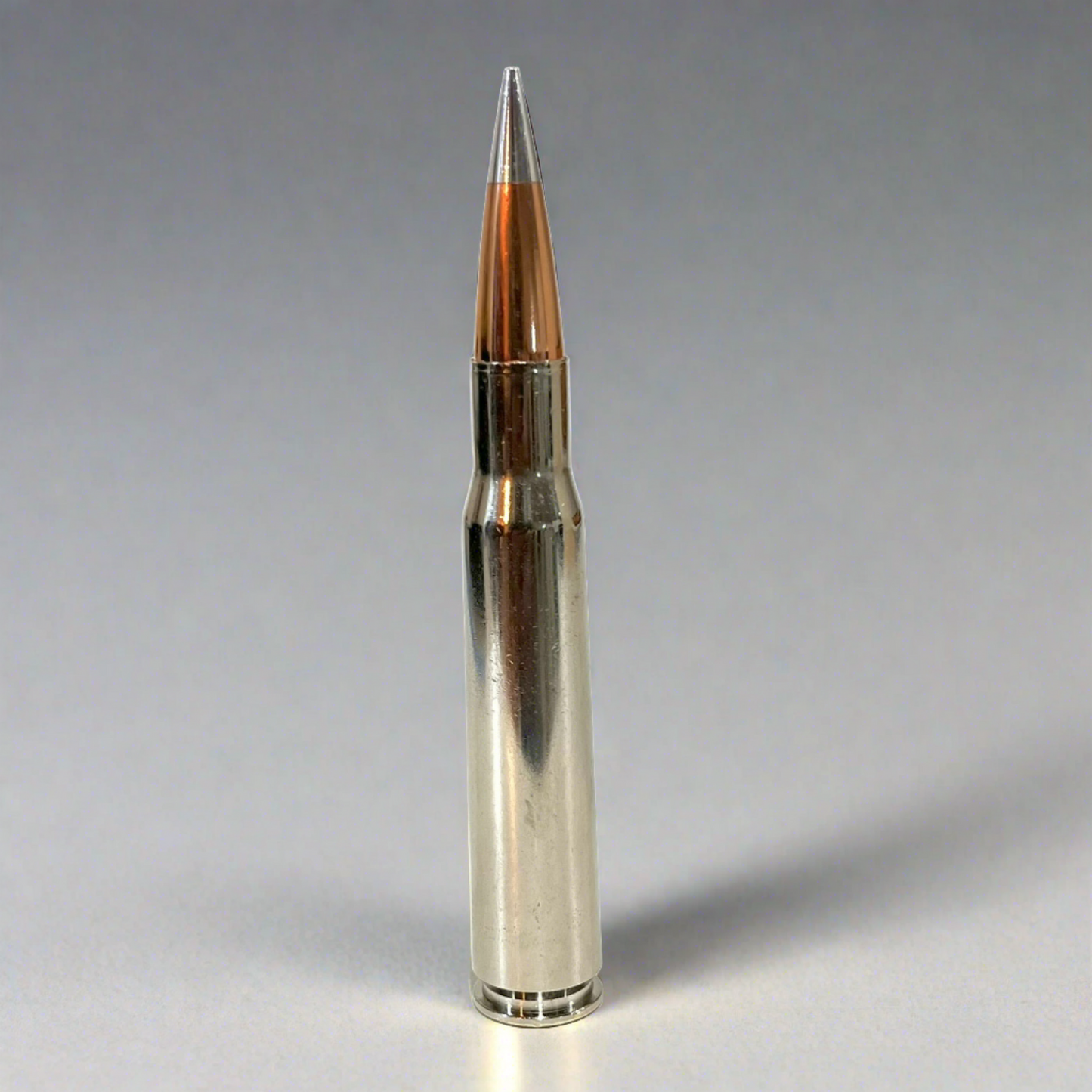 Polished silver nickel 50 cal BMG dummy round with copper bullet tip standing upright on reflective grey surface.
