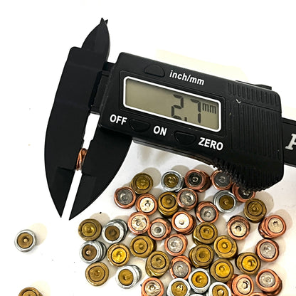 Digital caliper measuring the thickness of a spent primer at 2.7 mm over a cluster of mixed spent primers on a white background.
