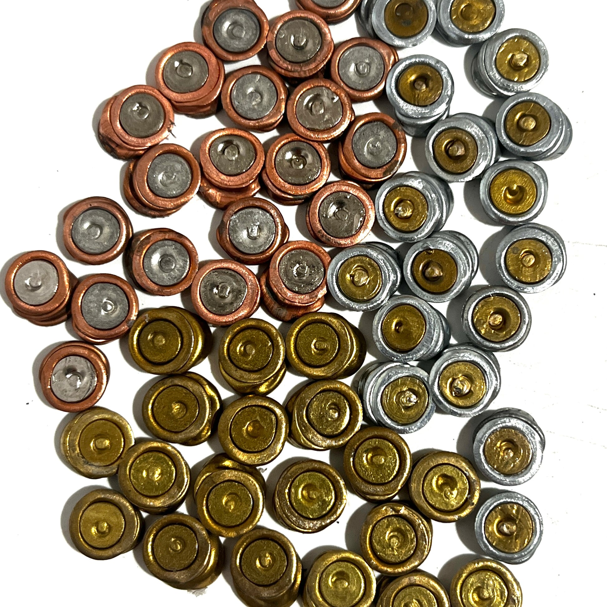 A group of spent primers flattened for earrings organized loosely by metallic colors including brass, copper, and silver on a white background.