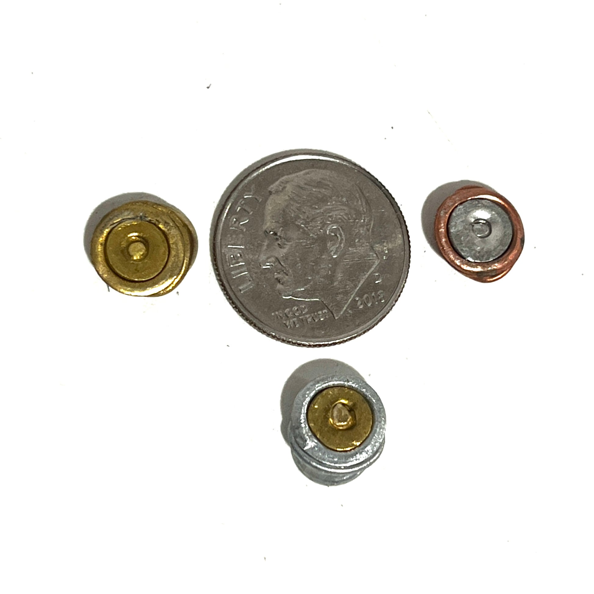 Three spent flattened primers of different metallic finishes shown next to a U.S. quarter for size reference on a white background.