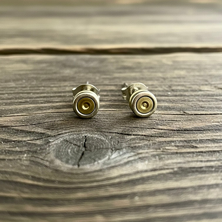 Two spent primers styled as stud earrings standing upright on a weathered grey wood surface.