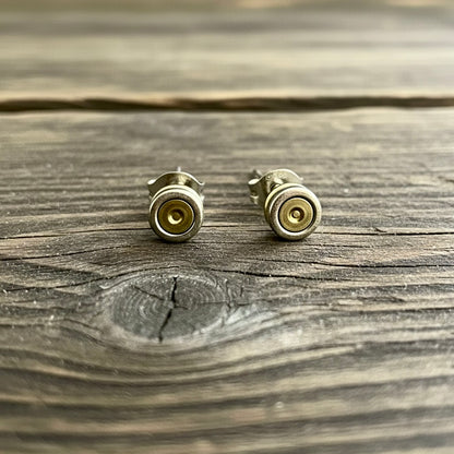 Two spent primers styled as stud earrings standing upright on a weathered grey wood surface.