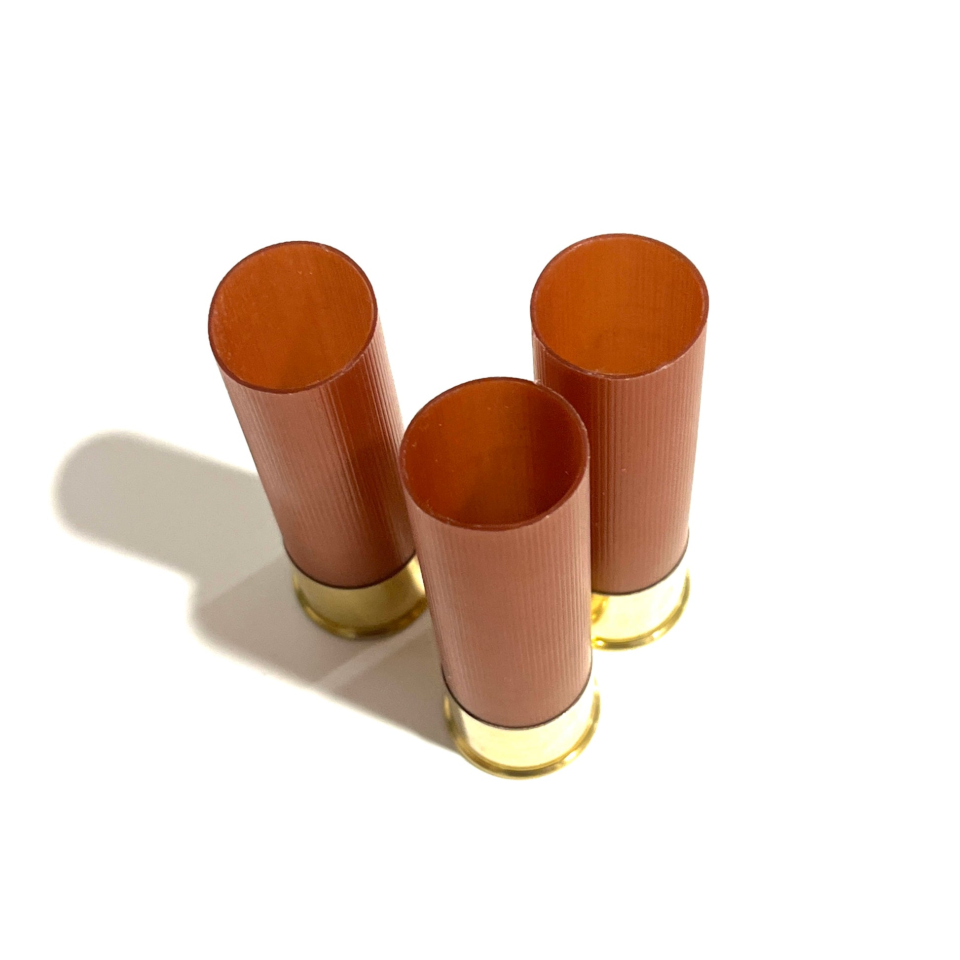 Three standing unmarked rose gold 12 gauge shotgun shell hulls with high brass bases on white background.
