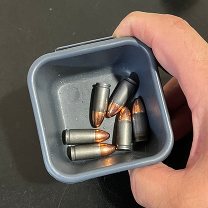 Five gray steel 9mm dummy cartridges held in a small blue tin container by hand