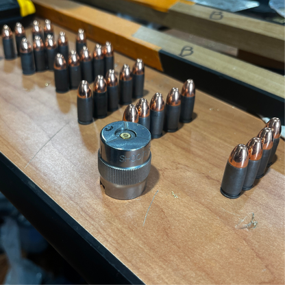 Numerous gray steel 9mm dummy cartridges standing in rows on a wooden workbench with a metal tool