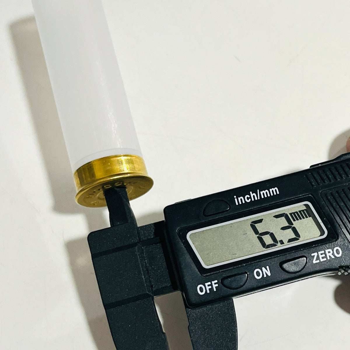 Digital caliper reading 6.3 mm with its jaw at the brass head primer pocket of a translucent, empty 12 gauge casing