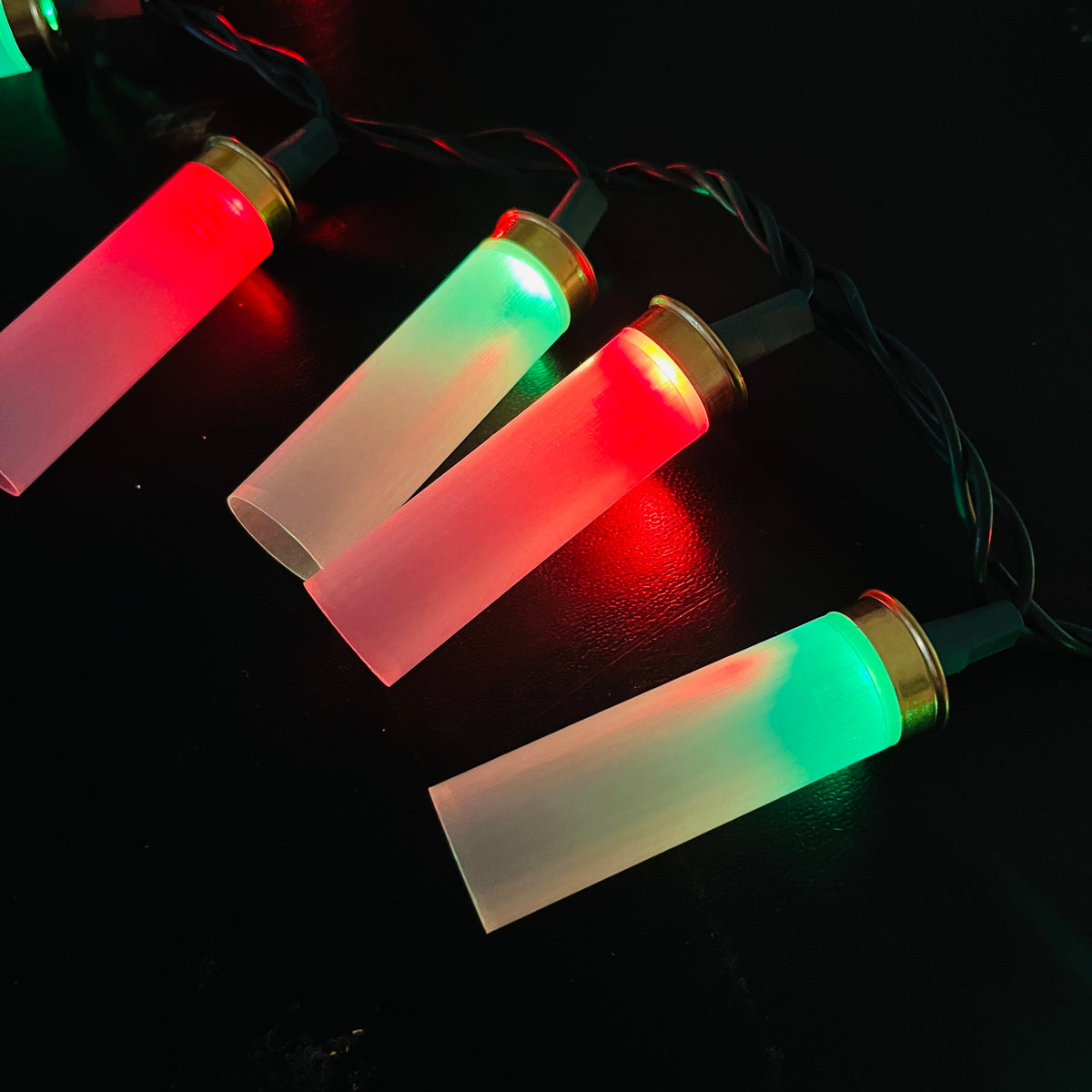 Four translucent empty shotgun shells on a Christmas light strand, glowing in alternating red and green on a dark surface