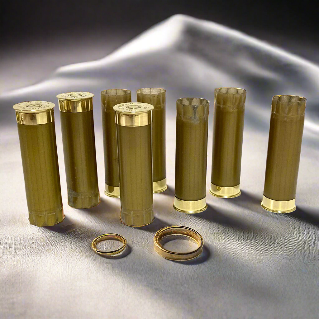 Eight empty gold shotgun casings standing upright on a white fabric surface with two rings in the foreground.