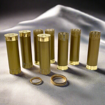 Eight empty gold shotgun casings standing upright on a white fabric surface with two rings in the foreground.