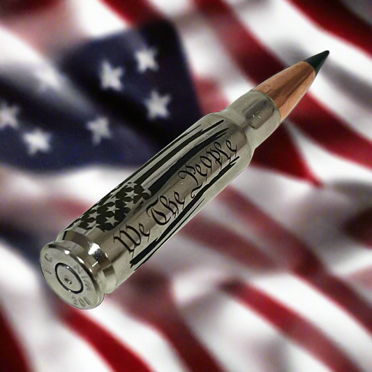 Nickel .308 rifle round with black polymer tip and We The People flag engraving, laid diagonally over blurred American flag background