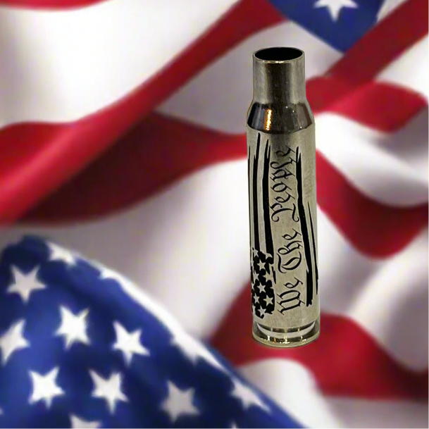 Nickel .308 WIN cartridge engraved with We The People, in front of a blurred American flag