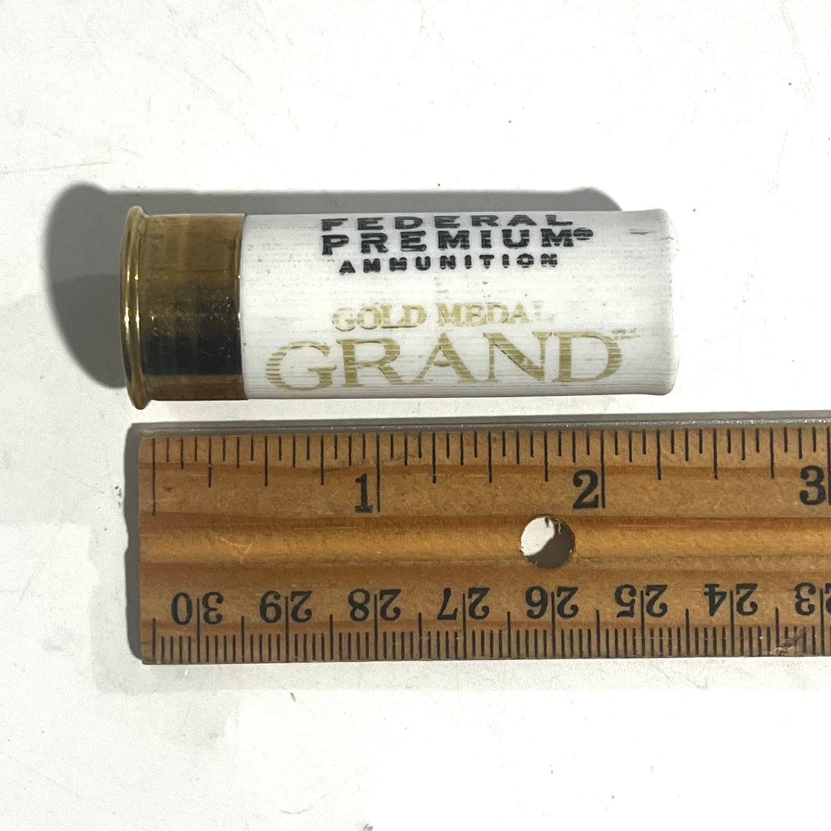 White Federal Premium Gold Medal Grand 12 gauge dummy shotgun shell laid next to a wooden ruler showing a length of about 2.75 inches