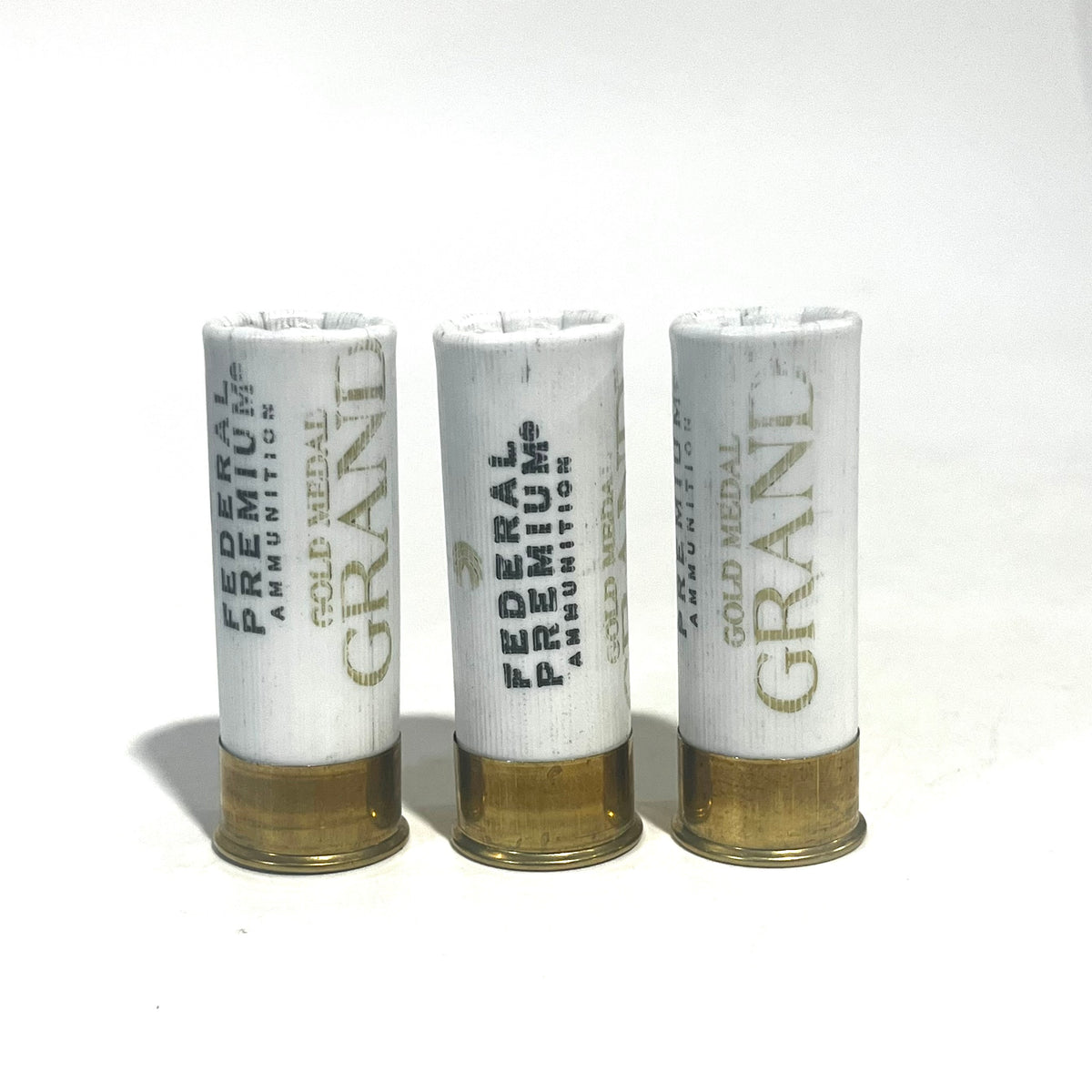 Three white Federal Premium Gold Medal Grand 12 gauge dummy shotgun shells with gold bases standing upright on a white background