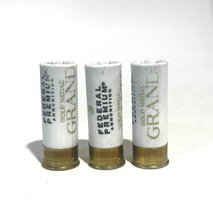 Three white Federal Premium Gold Medal Grand 12 gauge dummy shotgun shells with gold bases standing upright on a white background