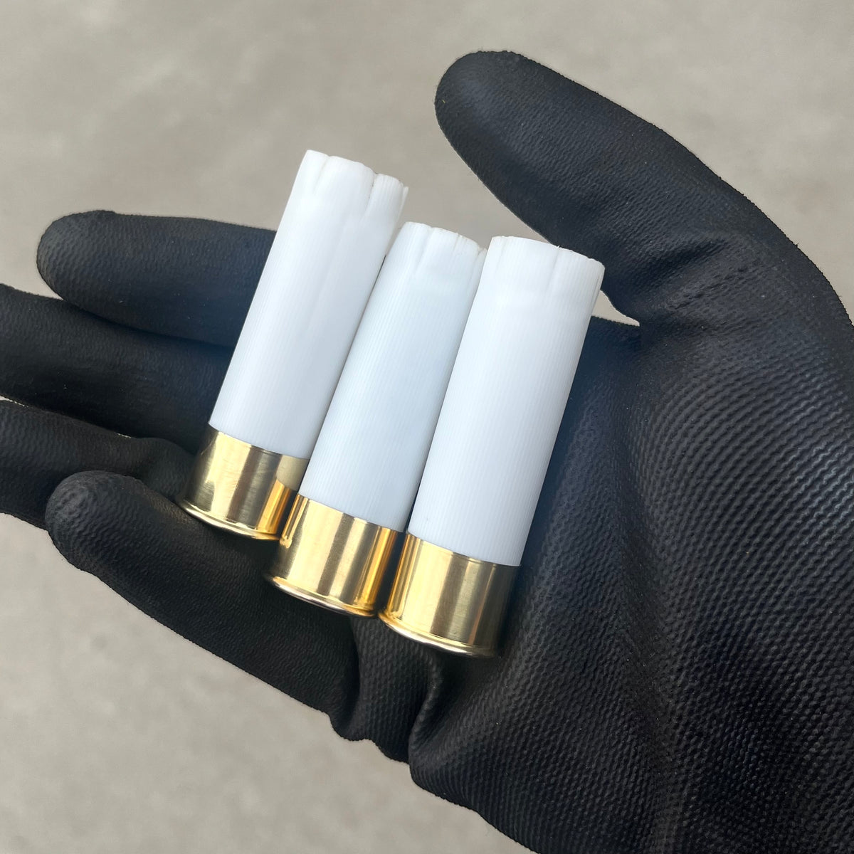 Gloved hand holding three white 12 gauge blank shotgun shells with brass bases