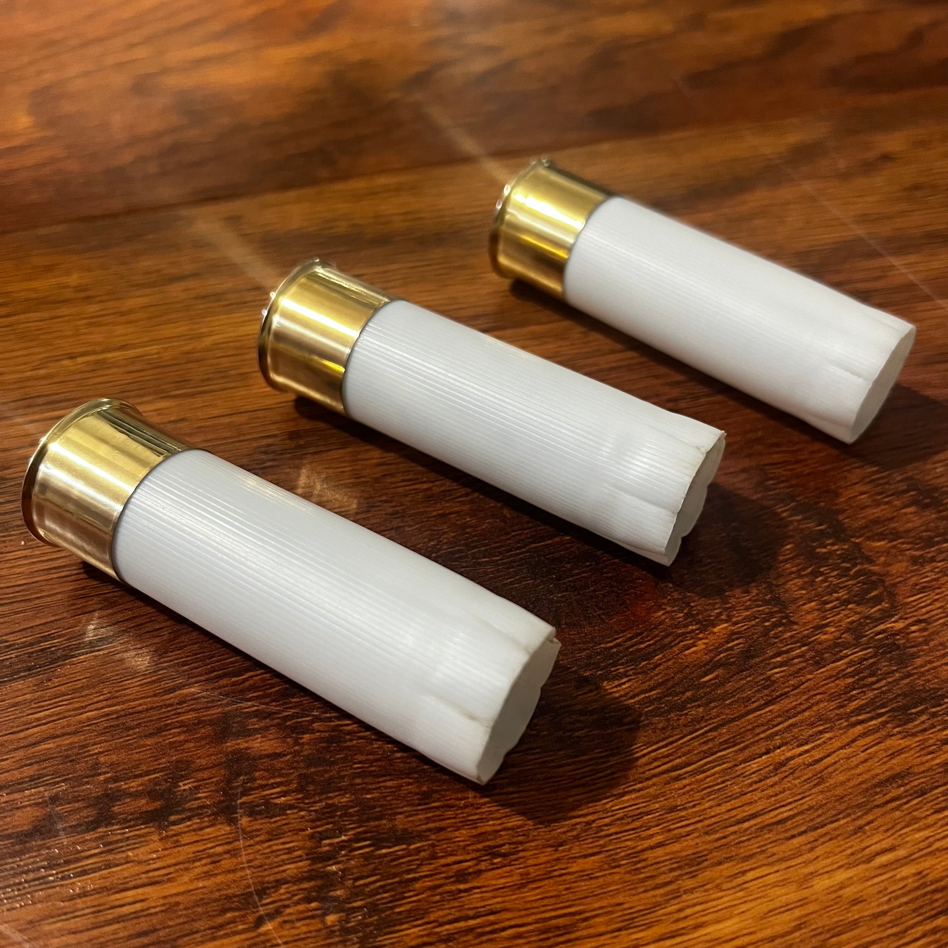 Three white 12 gauge blank shotgun shells with brass bases lying on a wood table