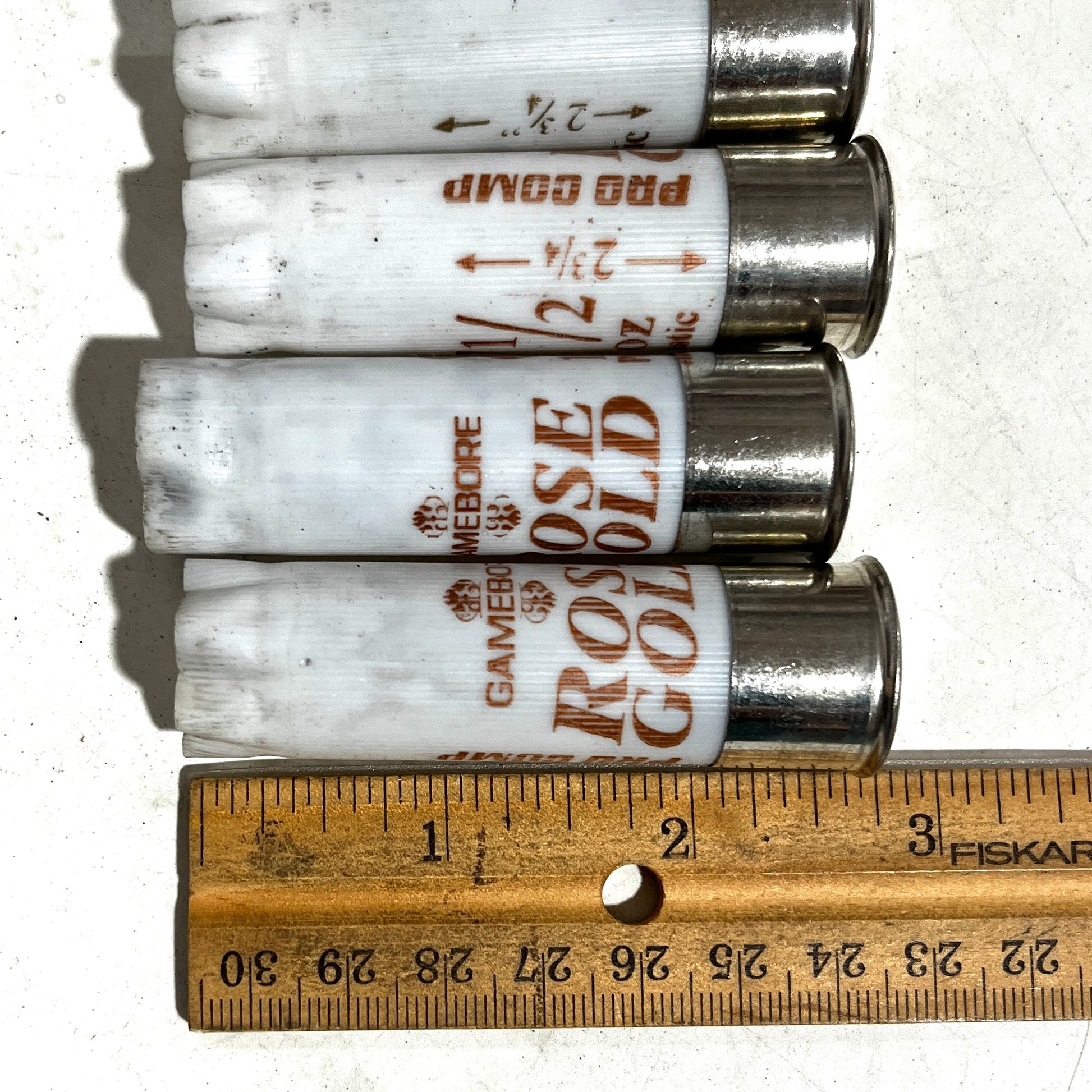 Three white Gamebore 12 gauge empty shotgun hullsdisplayed horizontally next to a wooden Fiskars ruler to show their 2-3/4 inch length.
