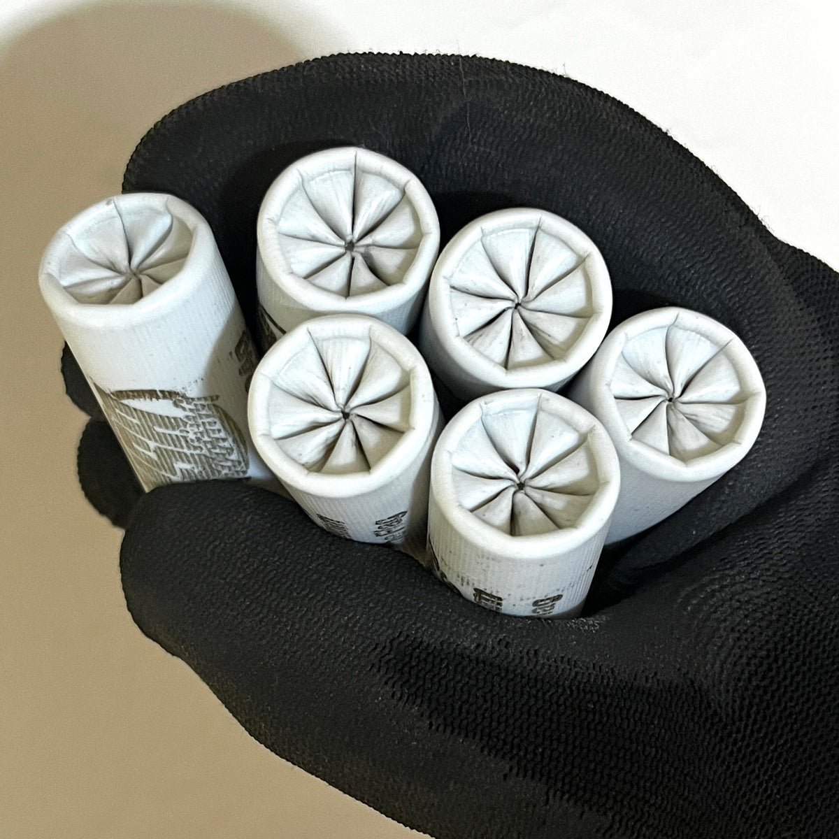 Six white spent shotgun shells with star-crimped tops held in a black gloved hand against a neutral background.