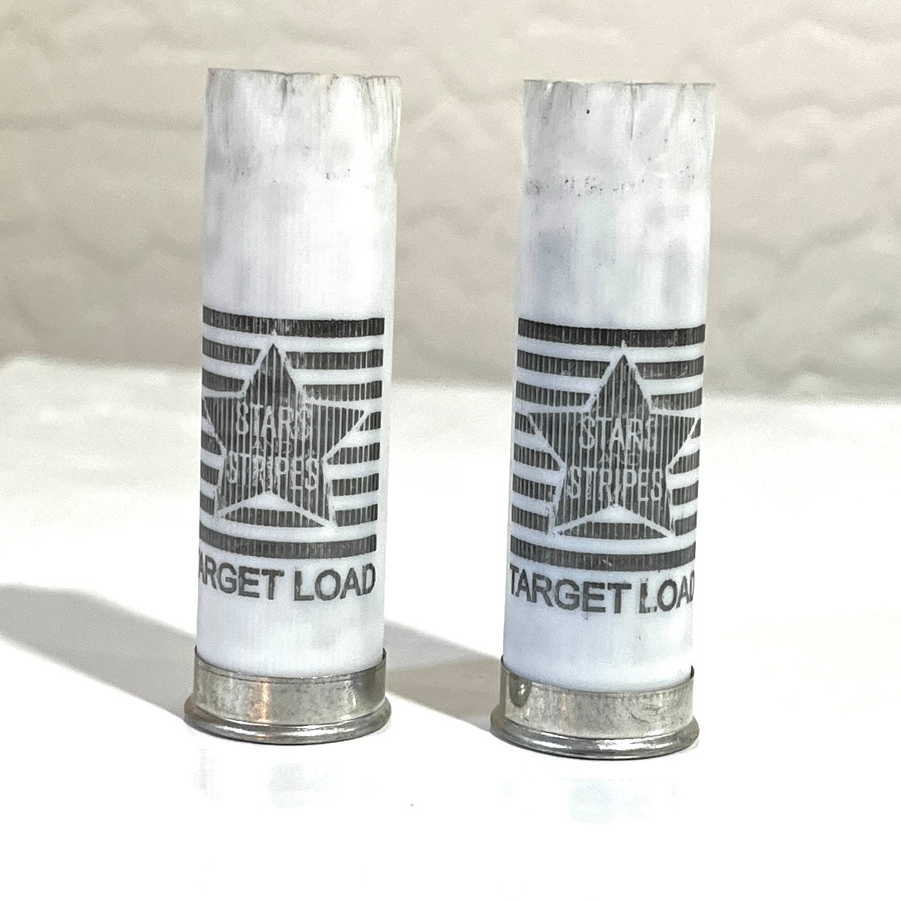 Two white 12 gauge empty shotgun shells printed Stars and Stripes Target Load with silver bases standing on a tile surface