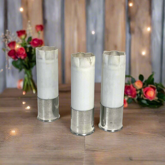 3 white shotgun shell hulls with silver high brass bases and crimped star-fold tops, standing on a wood table with roses and string lights