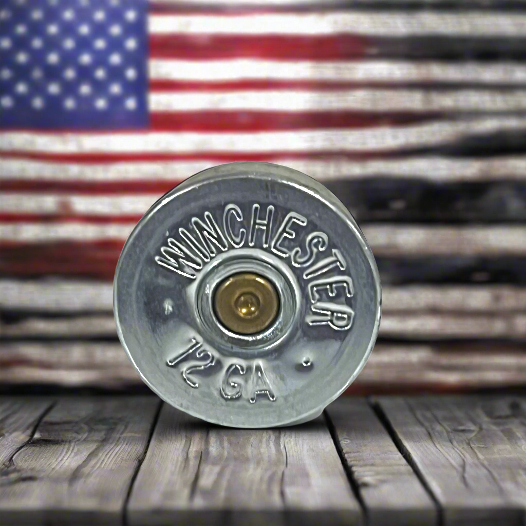 Single silver Winchester 12 gauge steel headstamp standing upright on a wood-plank surface, with a blurred American flag background, showing gold primer and stamped ‘Winchester 12 GA’ text