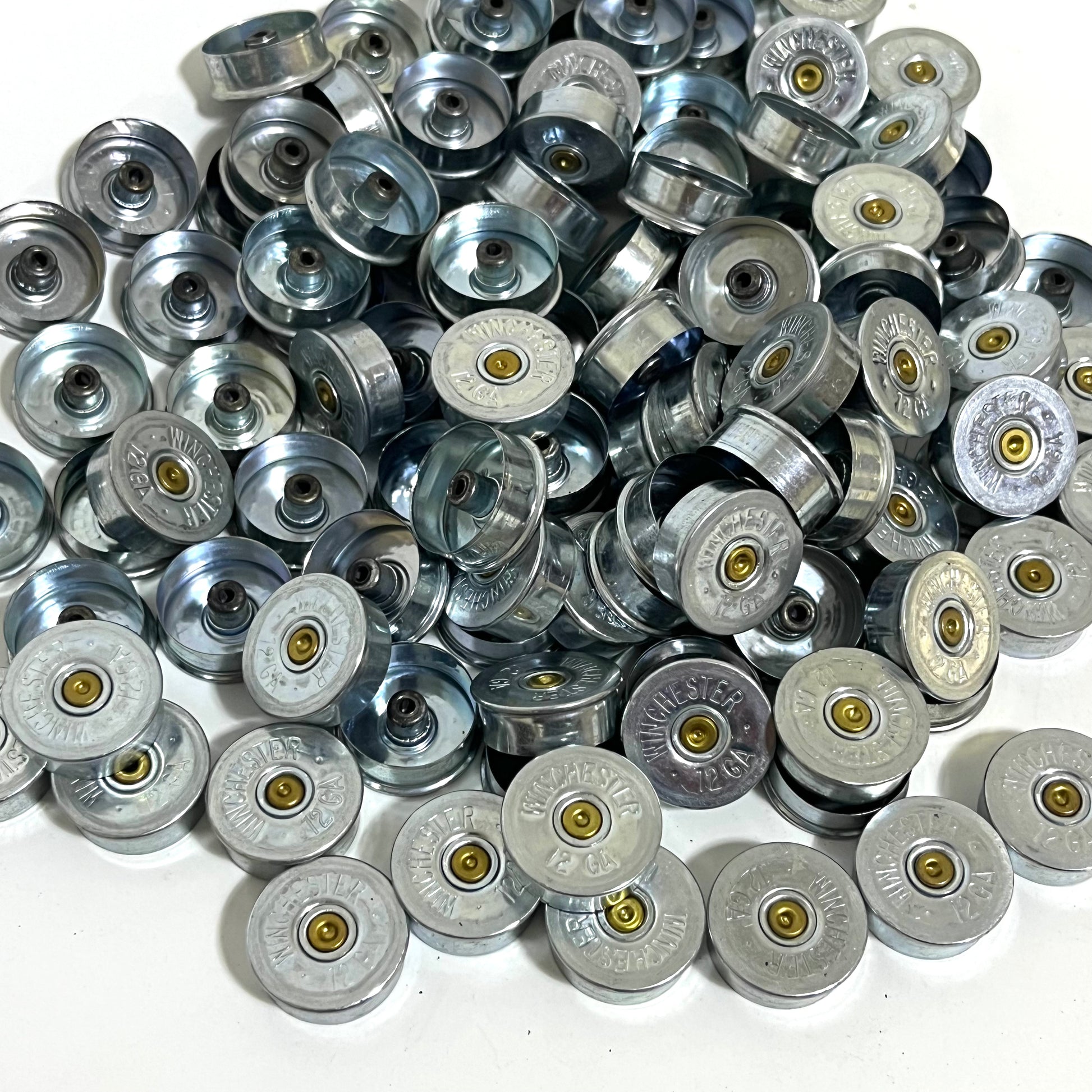 Large bulk pile of silver Winchester 12 gauge steel headstamps on a white background, showing gold primers and ‘Winchester 12 GA’ stampings