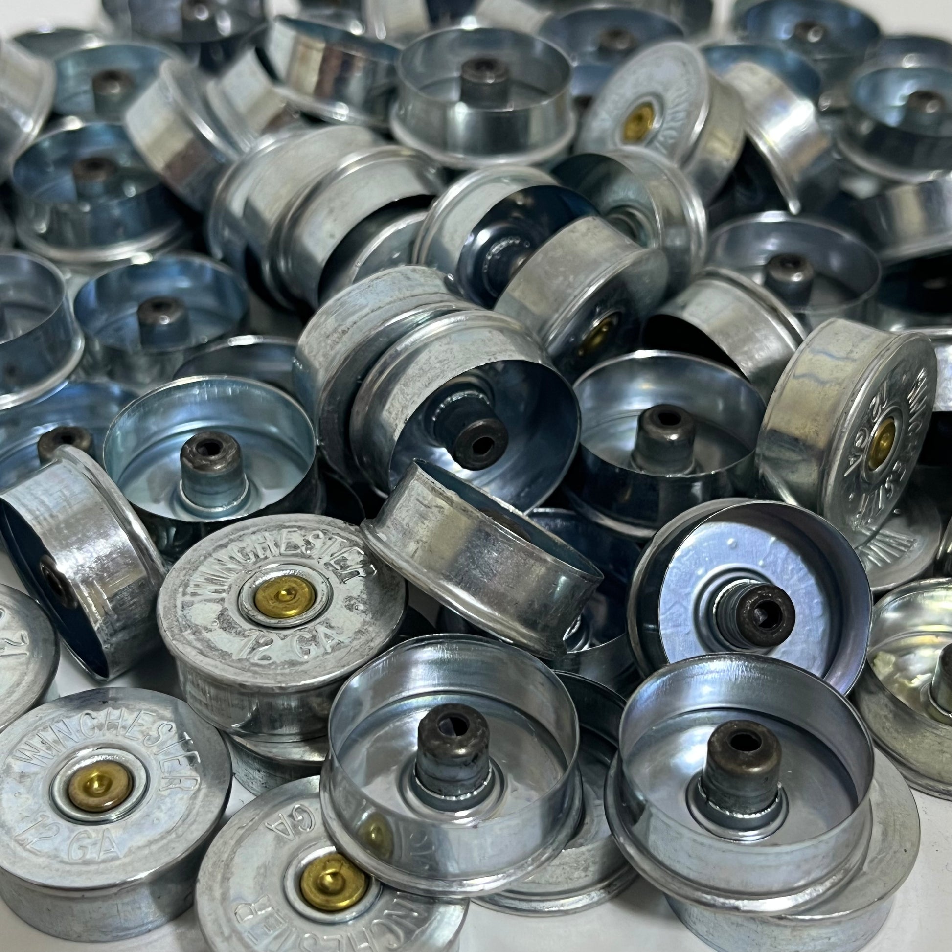 Large bulk pile of silver Winchester 12 gauge steel headstamps viewed at an angle, showing gold primers and hollow interiors
