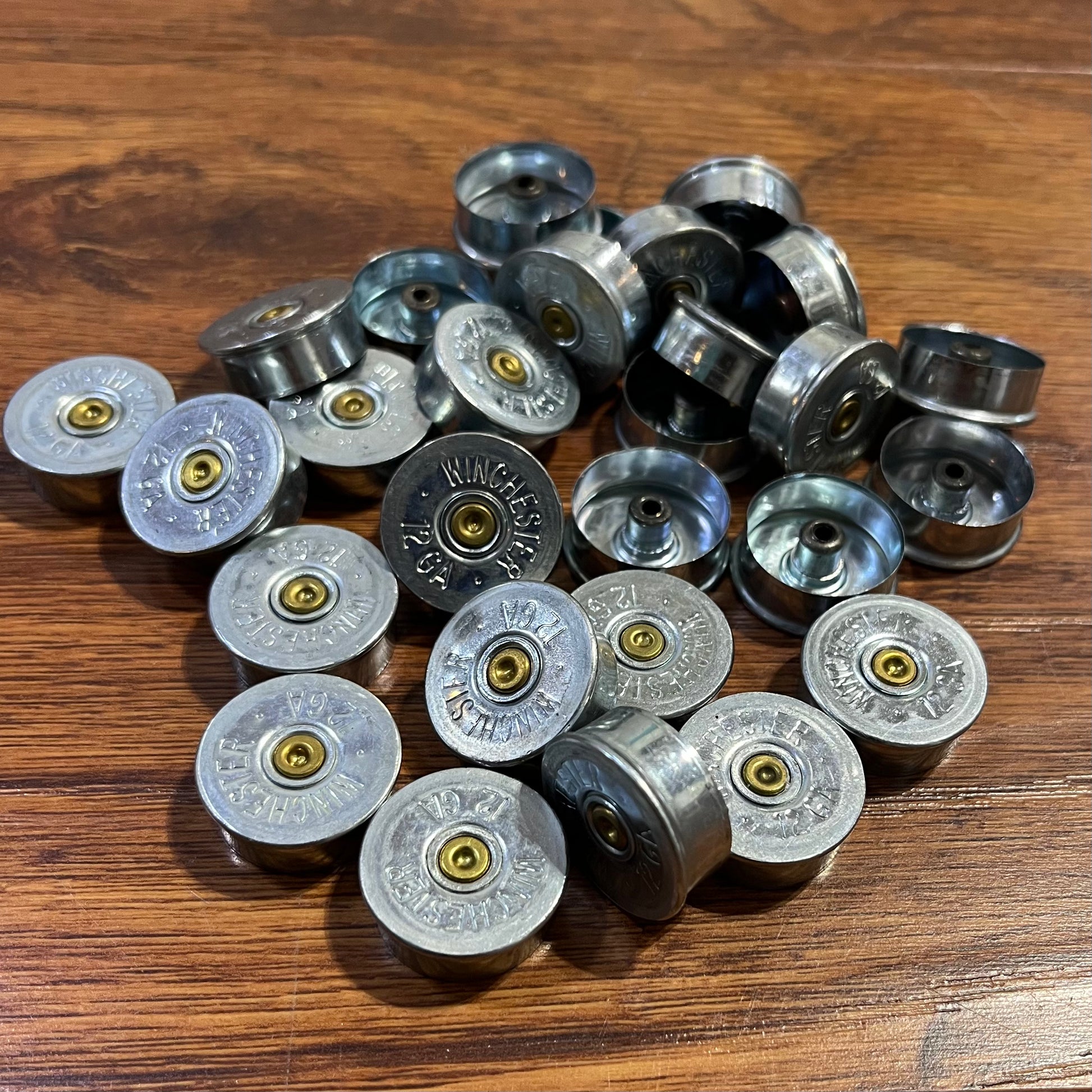 Bulk pile of silver Winchester 12 gauge steel headstamps on a wood-plank surface, showing gold primers and stamped ‘Winchester 12 GA’ text