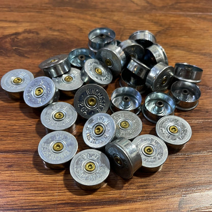 Bulk pile of silver Winchester 12 gauge steel headstamps on a wood-plank surface, showing gold primers and stamped ‘Winchester 12 GA’ text