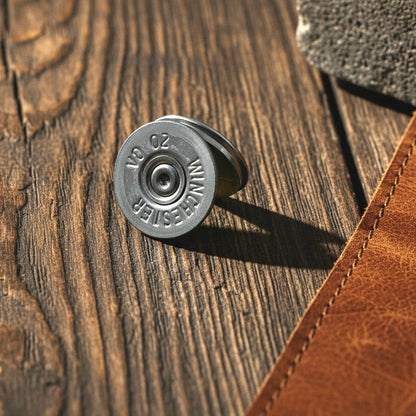 Silver 20 gauge Winchester shotgun shell slice with a visible primer resting on a wood plank next to a leather item