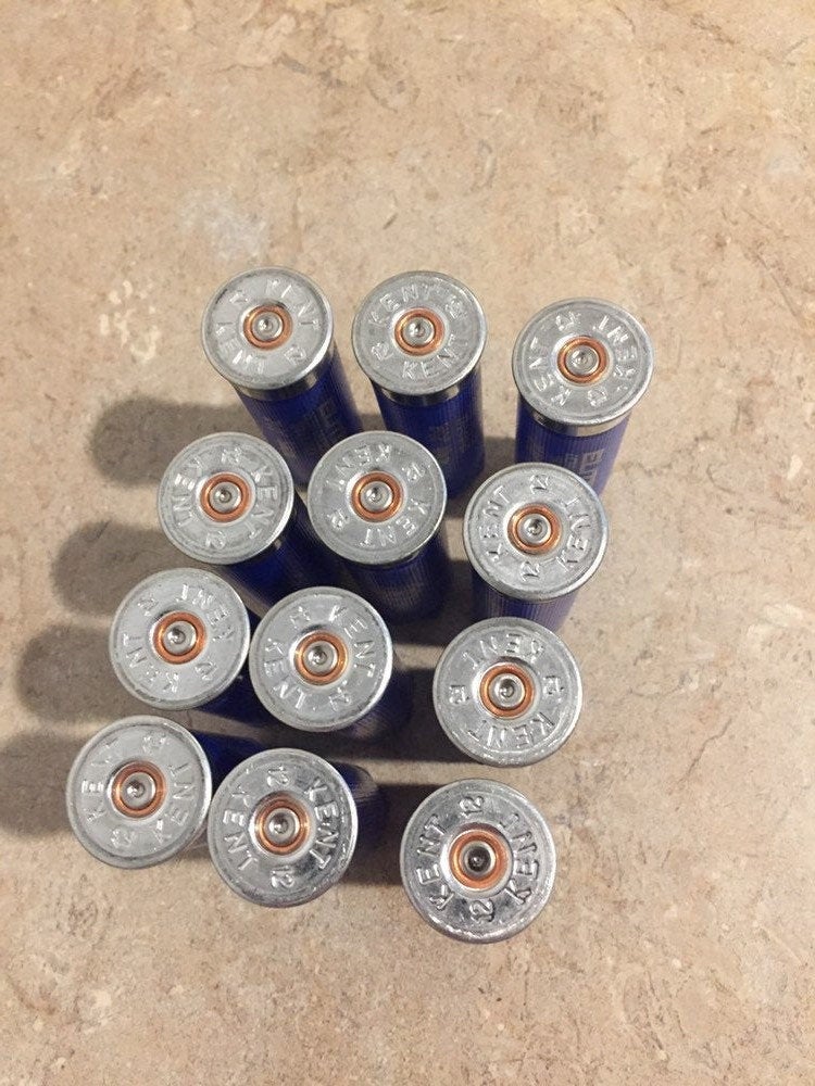 Group of 12 blue Kent 12-gauge empty shotgun shells or spent casings lying upside down on a textured surface showing silver metal bases with primer dimples.
