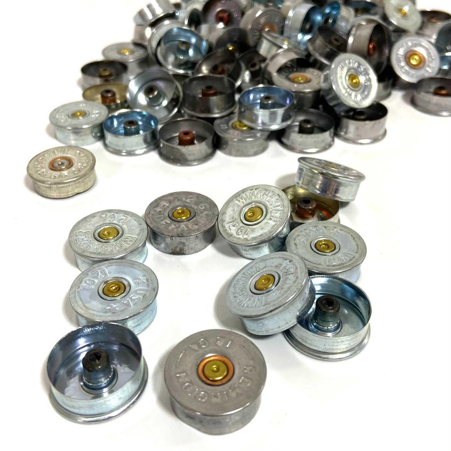 Silver 12 gauge shotgun shell headstamps in a close cluster with more piled behind on white background.