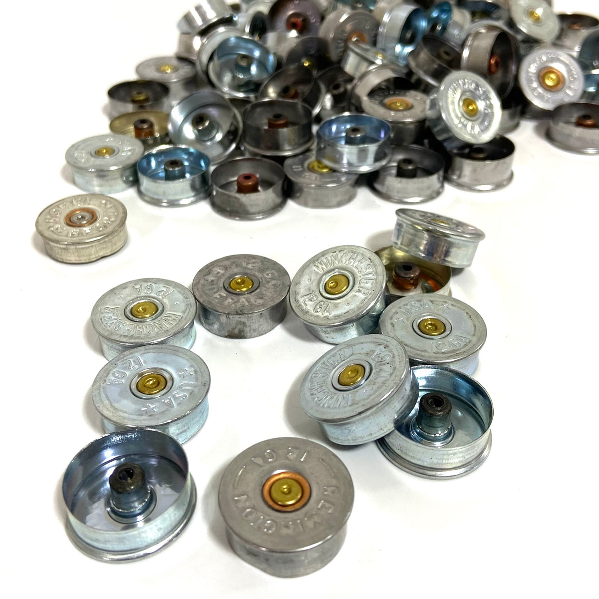 Silver 12 gauge shotgun shell headstamps in a close cluster with more piled behind on white background.