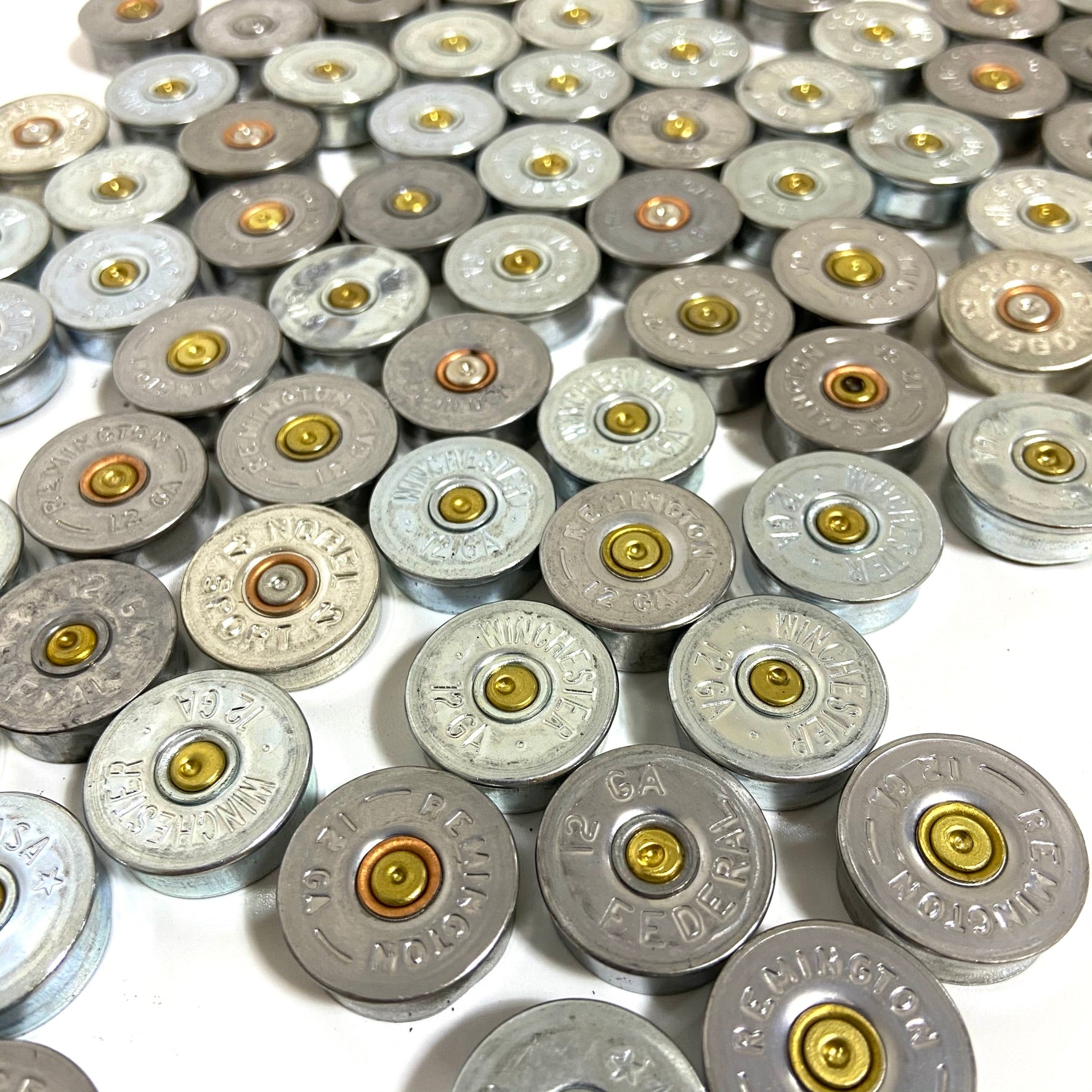 Silver 12 gauge shotgun shell headstamps packed together filling the frame, mixed manufacturer stamps.