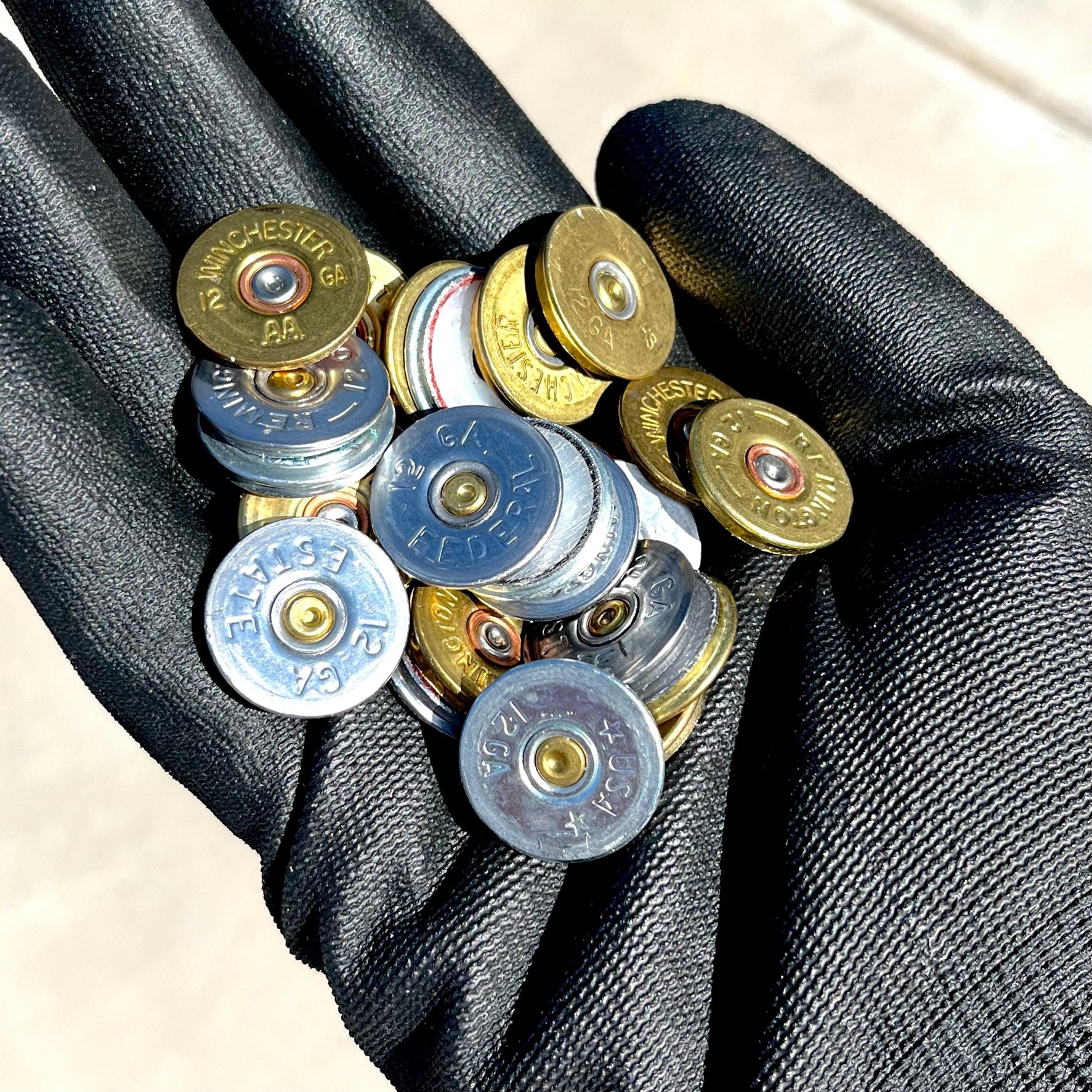 Black gloved hand holding gold and silver 12 gauge shotgun shell headstamp slices showing stamped brand markings.