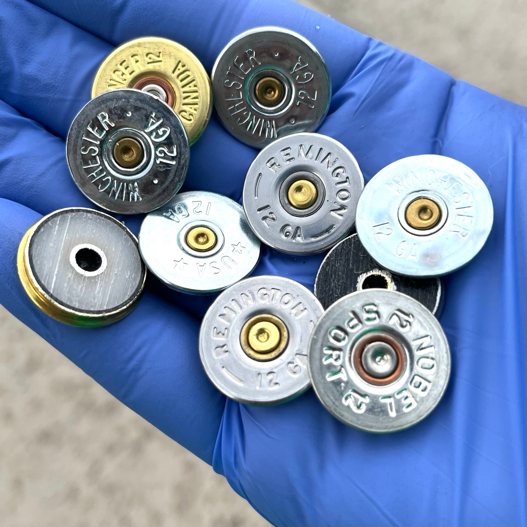 Blue gloved hand holding nine gold and silver 12 gauge shotgun shell headstamp slices showing stamped brand markings.