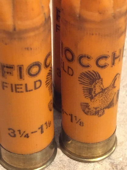 Two orange 12 gauge empty hulls showing Fiocchi Field and Gold printed text and pheasant graphic on stone surface.