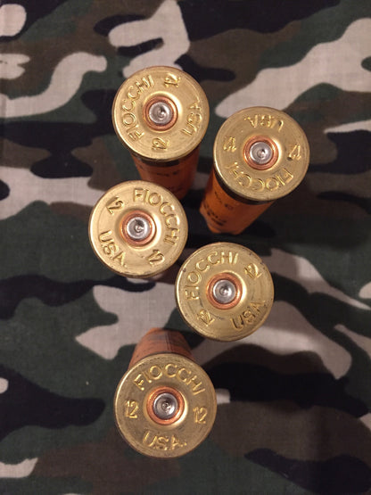 Five orange 12 gauge shotshell bases on camouflage fabric showing FIOCCHI 12 USA headstamps facing upward.